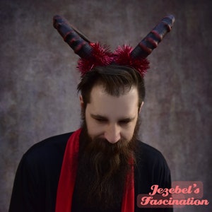 May include: A man with a long beard is wearing a red scarf and a red and black devil horn headband with red glitter. The text "Jezebel's Fascination" is visible in the lower right corner of the image.