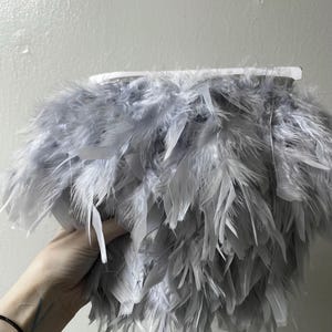 May include: A fluffy, gray feather boa. The boa is made of soft, light gray feathers attached to a white base. The feathers are layered, creating a full and voluminous appearance. The boa is held up against a white wall.