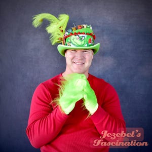 May include: A green top hat with a Grinch face and feather accents, paired with matching green gloves. The hat is decorated with red and white accents. The person is wearing a red long-sleeve shirt. The image is set against a dark blue background.
