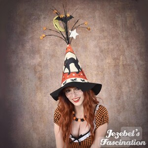 May include: A Halloween witch hat with a black cat silhouette, stars, and a crescent moon. The hat is orange, black, and white, with a wide brim. The witch is wearing an orange and black checkered dress.
