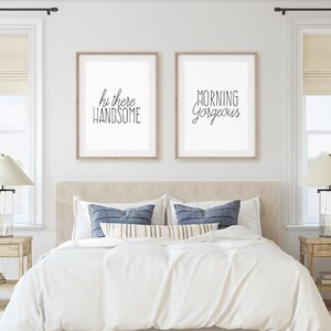 May include: Two framed prints with white mats and black text on a white background. The print on the left says "hi there handsome" and the print on the right says "morning gorgeous". The prints are hanging on a white wall above a bed with white bedding and blue pillows.
