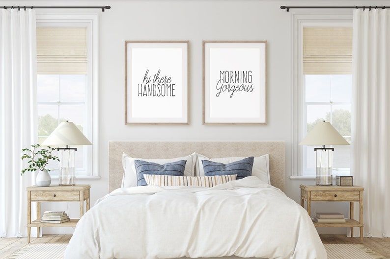 May include: Two framed prints with white mats and black text on a white background. The print on the left says "hi there handsome" and the print on the right says "morning gorgeous". The prints are hanging on a white wall above a bed with white bedding and blue pillows.