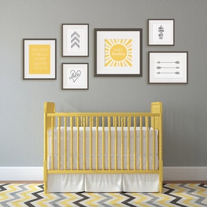 Puede incluir: Una cuna amarilla con ropa de cama blanca y una pared gris con impresiones enmarcadas en negro que representan flechas, un sol y el alfabeto. Las impresiones tienen texto amarillo.