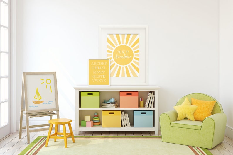 Puede incluir: Una estanter&iacute;a blanca con cestas de almacenamiento de color amarillo, verde, azul y naranja. La estanter&iacute;a est&aacute; decorada con dos impresiones enmarcadas, una con el alfabeto y la otra con un sol amarillo y el texto "You are my sunshine". Un sill&oacute;n verde con almohadas amarillas se encuentra frente a la estanter&iacute;a. Un taburete amarillo se encuentra frente a un caballete blanco con un dibujo de un barco y el sol.