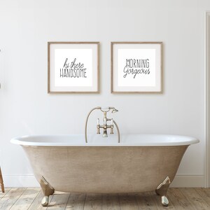 May include: A white bathroom with a freestanding bathtub, a wooden stool, and two framed prints on the wall. The prints say "Hi there handsome" and "Morning Gorgeous".