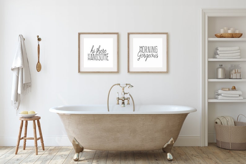 May include: A white bathroom with a freestanding bathtub, a wooden stool, and two framed prints on the wall. The prints say "Hi there handsome" and "Morning Gorgeous".