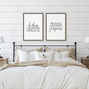 May include: Two framed prints with white backgrounds and black text on a white shiplap wall. The text on the left print reads "hi there handsome" and the text on the right print reads "morning gorgeous". The prints are above a white bed with a beige duvet and a beige throw blanket. There are two nightstands with lamps and plants on either side of the bed.