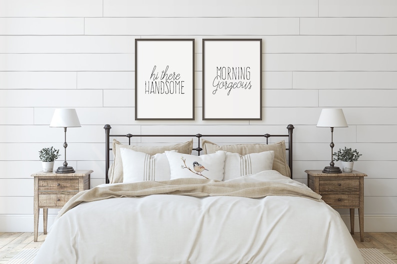 May include: Two framed prints with white backgrounds and black text on a white shiplap wall. The text on the left print reads "hi there handsome" and the text on the right print reads "morning gorgeous". The prints are above a white bed with a beige duvet and a beige throw blanket. There are two nightstands with lamps and plants on either side of the bed.