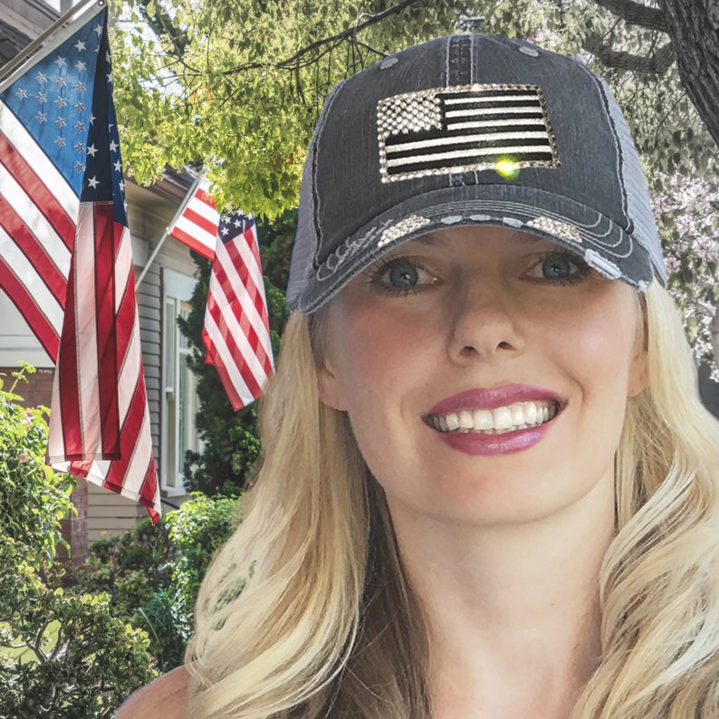 May include: A woman wearing a black and grey distressed baseball cap with a rhinestone American flag design. The cap has a mesh back.