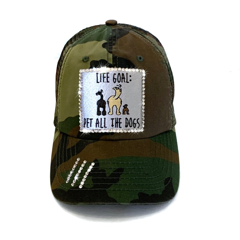 May include: A camouflage trucker hat with a white patch that says "Life Goal: Pet All The Dogs" with a cartoon image of three dogs. The patch is decorated with rhinestones.