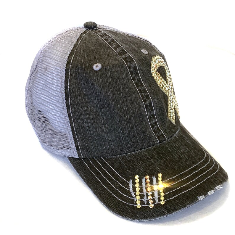 May include: A distressed black denim baseball cap with a gray mesh back. The cap features a sparkling gold ribbon design on the front and a small cluster of gold rhinestones near the brim. The brim has a distressed, frayed edge.
