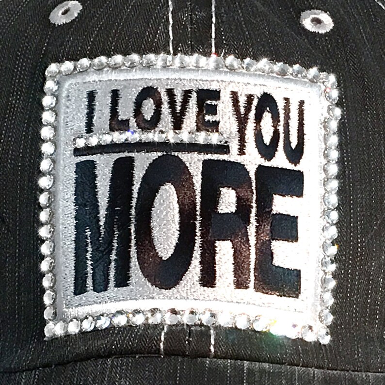 K&ouml;nnte beinhalten: Schwarze Baseballkappe mit einem silbernen Aufn&auml;her mit dem schwarzen Schriftzug "I LOVE YOU MORE". Der Aufn&auml;her ist mit Strasssteinen umrandet. Die Kappe hat ein schwarz-wei&szlig;es Nadelstreifenmuster.