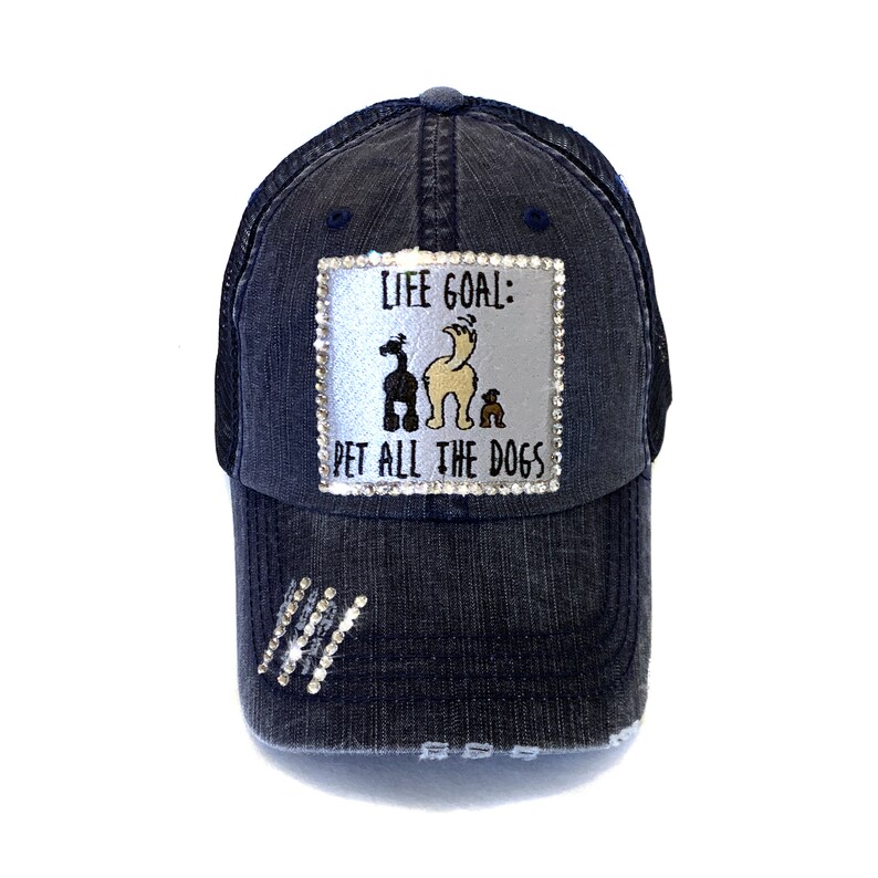 May include: A dark blue denim trucker hat with a silver patch that reads "Life Goal: Pet All The Dogs". The patch features a cartoon image of three dogs. The hat has a mesh back and a distressed look.