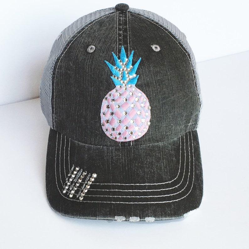 May include: Black denim baseball cap with a gray mesh back. The cap features a pink and silver rhinestone pineapple design on the front. Additional rhinestones are arranged on the brim. A stylish accessory.