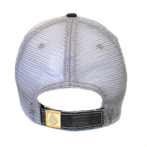 May include: A gray trucker hat with a mesh back and a dark gray brim. The hat features a small, dark gray button on top and a gold-colored patch on the adjustable strap.