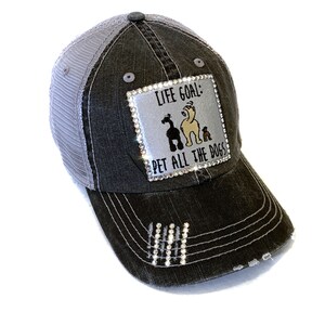 May include: A black and gray distressed trucker hat with a silver patch that reads "Life Goal: Pet All The Dogs" with a rhinestone border and a cartoon image of three dogs. The hat has a mesh back and rhinestones on the bill.