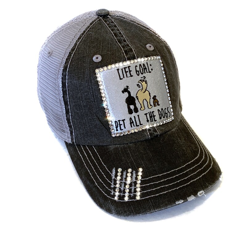 May include: A black and gray distressed trucker hat with a silver patch that reads "Life Goal: Pet All The Dogs" with a rhinestone border and a cartoon image of three dogs. The hat has a mesh back and rhinestones on the bill.
