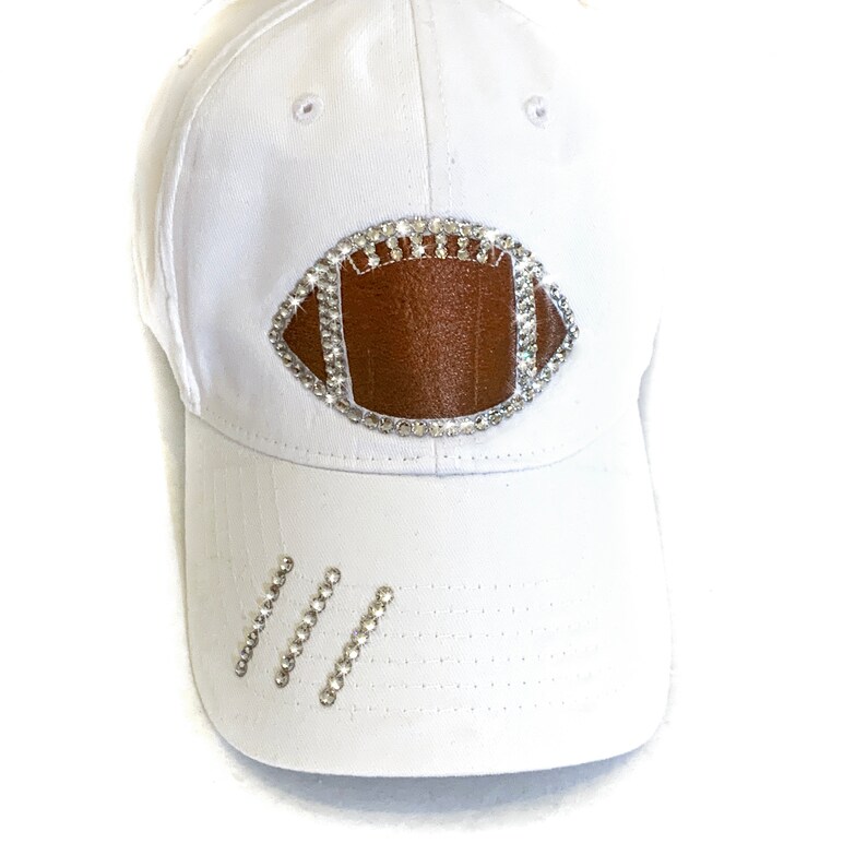 May include: White baseball cap with a brown football design made of rhinestones. The football has a white outline and is centered on the front of the cap.