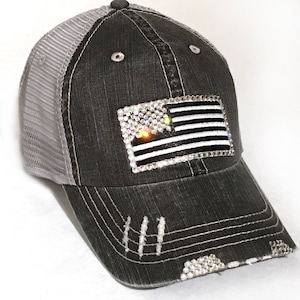 May include: A black and gray distressed trucker hat with a mesh back. The hat features a rhinestone American flag design.