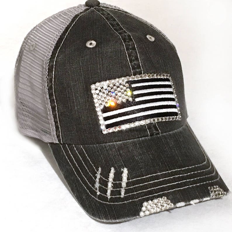 May include: A black and gray distressed trucker hat with a mesh back. The hat features a rhinestone American flag design.