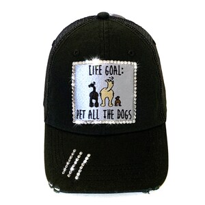 May include: Black trucker hat with a white patch that says "Life Goal: Pet All The Dogs" with a cartoon image of three dogs. The patch is decorated with rhinestones.