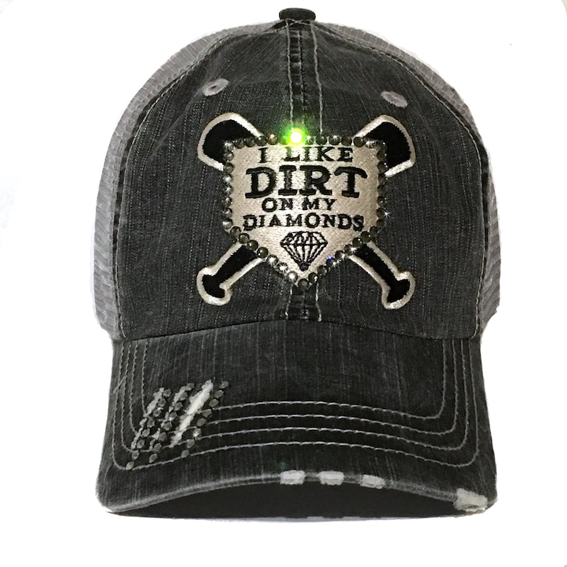 May include: A black and gray baseball cap with a white diamond-shaped patch that says "I like dirt on my diamonds" with two crossed baseball bats. The patch has rhinestones.