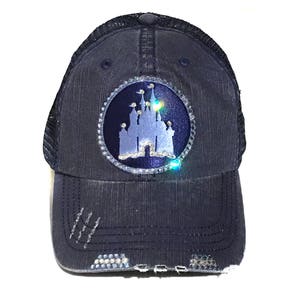 Puede incluir: Una gorra de camionero de mezclilla azul oscuro con un aspecto desgastado. La gorra presenta un parche redondo plateado con una silueta blanca de un castillo y diamantes de imitaci&oacute;n.