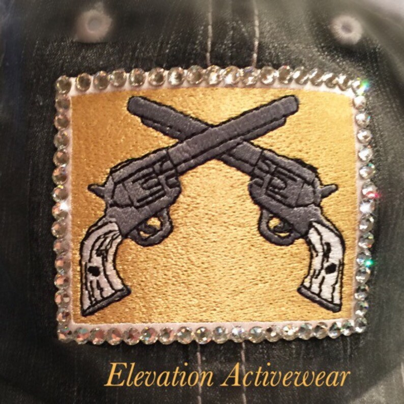 May include: A black denim baseball cap with a gold and gray embroidered patch of two crossed pistols. The patch is surrounded by a border of clear rhinestones. The text "Elevation Activewear" is printed below the patch.