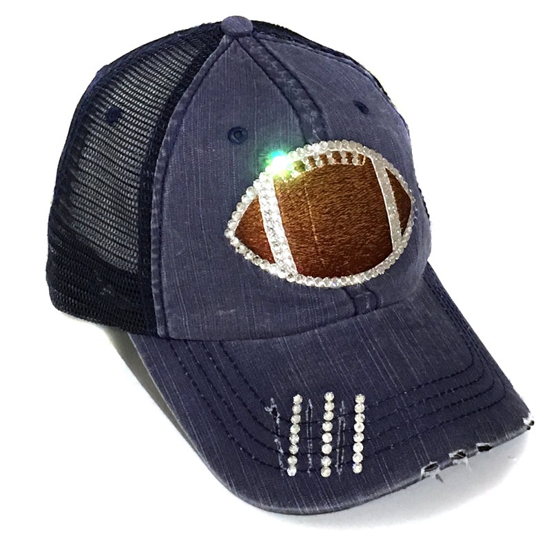 May include: A navy blue trucker hat with a distressed denim look. The hat features a rhinestone-encrusted football design on the front and rhinestones on the bill.
