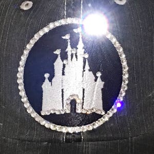 Puede incluir: Una imagen bordada en blanco y negro del castillo de Cenicienta rodeada de un c&iacute;rculo de diamantes de imitaci&oacute;n sobre una tela gris oscura.