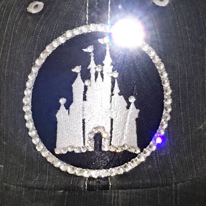 Puede incluir: Una imagen bordada en blanco y negro del castillo de Cenicienta rodeada de un c&iacute;rculo de diamantes de imitaci&oacute;n sobre una tela gris oscura.