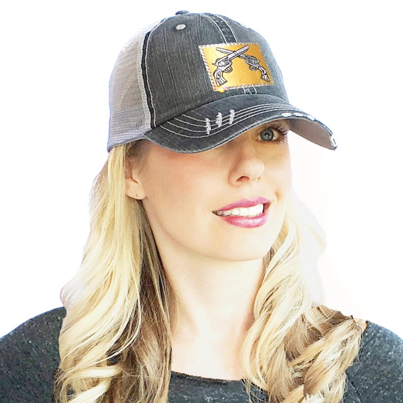 May include: A black and gray distressed trucker hat with a gold patch featuring a pair of crossed pistols.