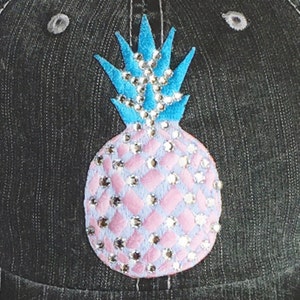 May include: A denim baseball cap featuring a bedazzled pineapple design. The pineapple has a light blue crown and a pink body with diamond-shaped accents, all embellished with sparkling rhinestones. The cap has a dark gray color.