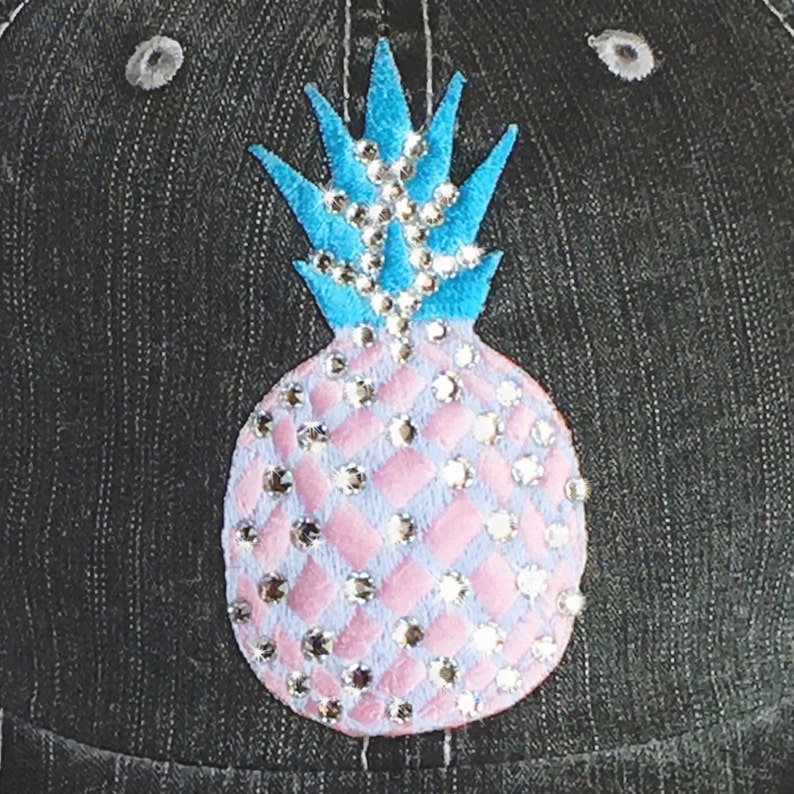 May include: A denim baseball cap featuring a bedazzled pineapple design. The pineapple has a light blue crown and a pink body with diamond-shaped accents, all embellished with sparkling rhinestones. The cap has a dark gray color.