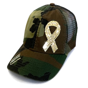 May include: Camouflage baseball cap with a mesh back and a sparkling, rhinestone-studded ribbon emblem. The cap features a curved brim with additional rhinestone accents. The color palette includes shades of green, brown, and black.