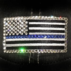 Puede incluir: Una bandera estadounidense en blanco y negro con una franja azul, hecha de diamantes de imitación. La bandera está sobre un fondo negro.