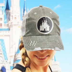Puede incluir: Una gorra de camionero gris y blanca con un parche negro y blanco de un castillo incrustado con diamantes de imitaci&oacute;n. La gorra la lleva puesta una persona con el pelo rubio y una sonrisa.