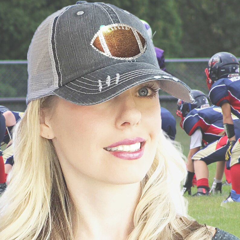 May include: A woman wearing a gray and white baseball cap with a rhinestone football design. The cap has a mesh back and a distressed look.