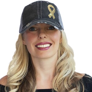May include: A distressed black trucker hat with a gold sequined ribbon design. The hat features a mesh back and a curved brim, with a worn, vintage aesthetic. The ribbon is a symbol of awareness.