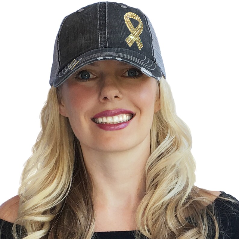 May include: A distressed black trucker hat with a gold sequined ribbon design. The hat features a mesh back and a curved brim, with a worn, vintage aesthetic. The ribbon is a symbol of awareness.