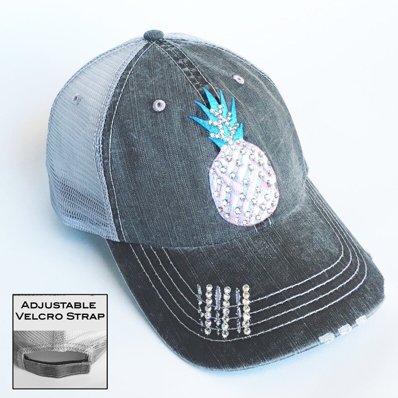 May include: Distressed charcoal gray baseball cap with a light gray mesh back. The cap features a bedazzled pineapple design with a blue crown and a white and pink body. The brim has decorative rhinestone accents and the text "ADJUSTABLE VELCRO STRAP".