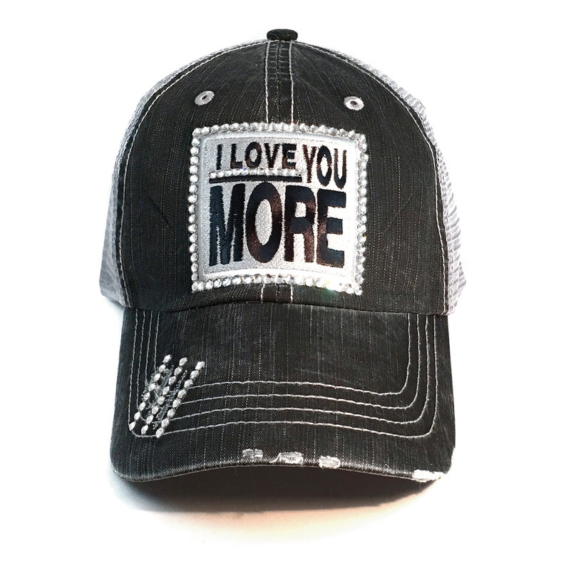 K&ouml;nnte beinhalten: Schwarze Denim-Baseballkappe mit wei&szlig;em Mesh-R&uuml;cken. Auf der Vorderseite steht in Schwarz und Silber der Schriftzug "I LOVE YOU MORE", umrahmt von Strasssteinen. Der Schirm hat dekorative N&auml;hte und Strass-Akzente.