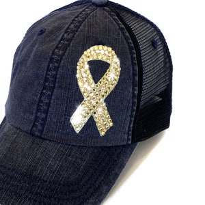May include: A navy blue baseball cap with a mesh back. The cap features a sparkling, rhinestone-covered ribbon emblem. The ribbon is a light gold color and is centered on the front of the cap.