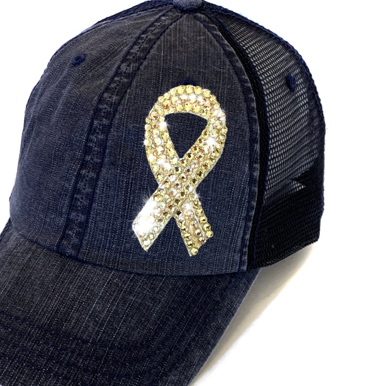 May include: A navy blue baseball cap with a mesh back. The cap features a sparkling, rhinestone-covered ribbon emblem. The ribbon is a light gold color and is centered on the front of the cap.