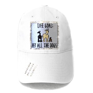 May include: White baseball cap with a rhinestone-encrusted patch that reads "Life Goal: Pet All The Dogs" and features a cartoon image of three dogs.