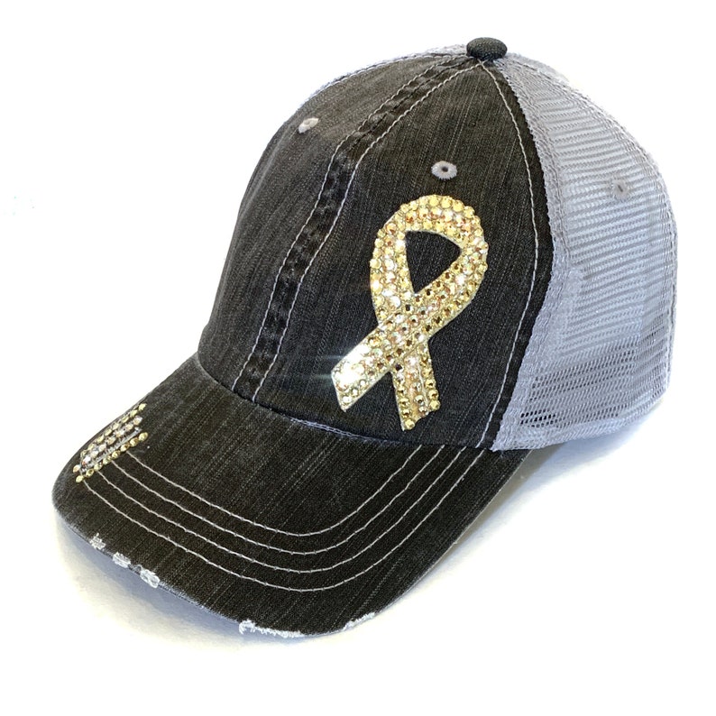 May include: A distressed black denim baseball cap with a gray mesh back. The cap features a gold rhinestone ribbon design on the front. The brim has a distressed edge with rhinestones.