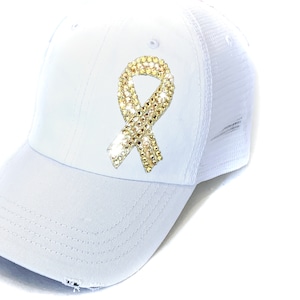 May include: White baseball cap with a mesh back and a curved brim. The cap features a gold rhinestone ribbon design on the front. The hat has a distressed look on the brim.