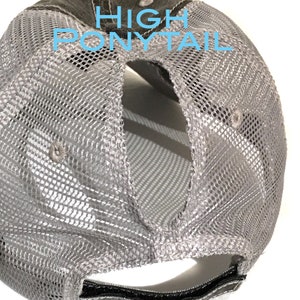 May include: A gray mesh trucker hat with a high ponytail opening. The hat features a camouflage pattern on the top and the text "HIGH PONYTAIL" in light blue. The back of the hat is made of a gray mesh material.