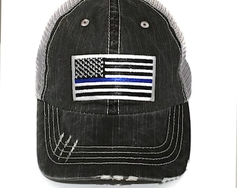 Puede incluir: Una gorra de camionero negra y gris desgastada con un parche de bandera estadounidense blanca y azul y una fina línea azul. La gorra tiene una parte trasera de malla.