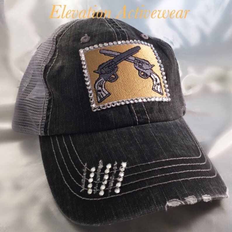 May include: A black and gray distressed trucker hat with a gray mesh back. The hat features a gold square patch with a rhinestone border and a design of two crossed pistols. The hat also has rhinestones on the brim.